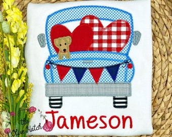 May include: White t-shirt featuring a blue gingham truck carrying red hearts and a small dog. The truck has a banner of red and blue pennants. The name "Jameson" is embroidered in red.