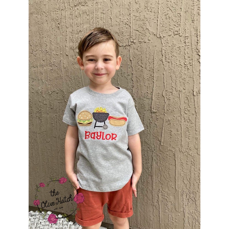 May include: A grey t-shirt with a red embroidered design of a grill, hamburger, hot dog, and flames. The text "Baylor" is embroidered in red below the design.