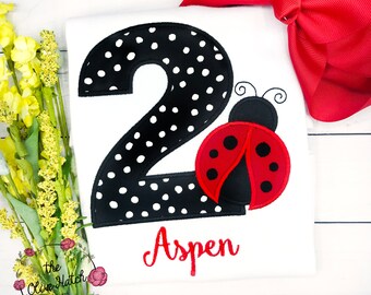 May include: A white t-shirt with a black and white polka dot number 2 and a red ladybug with black spots. The name Aspen is embroidered in red below the number.