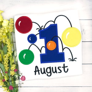 May include: A white t-shirt with a blue number one and the word "August" embroidered on it. The number one is surrounded by colorful bouncing balls.