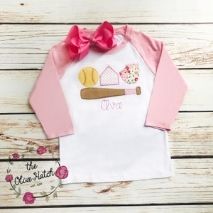 Puede incluir: Una camiseta de b&eacute;isbol de manga larga blanca y rosa con un lazo rosa en la parte superior. La camiseta tiene un dise&ntilde;o bordado de una pelota de b&eacute;isbol, un bate y un plato de home con el nombre "Ava" debajo.