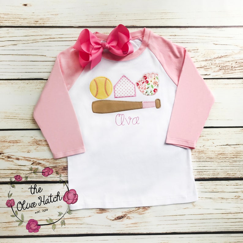 Puede incluir: Una camiseta de b&eacute;isbol de manga larga blanca y rosa con un lazo rosa en la parte superior. La camiseta tiene un dise&ntilde;o bordado de una pelota de b&eacute;isbol, un bate y un plato de home con el nombre "Ava" debajo.