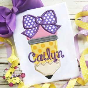 May include: White t-shirt featuring a pencil design with a purple bow and the name "Cailyn" in purple script. The pencil has a yellow polka-dot pattern and a light pink eraser. A large purple bow is at the top left.
