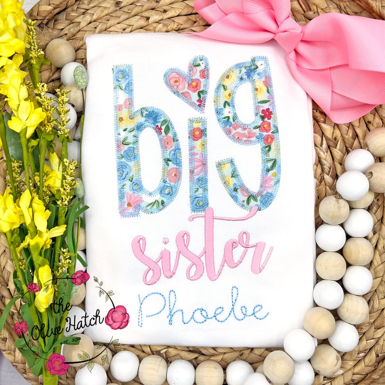 May include: White onesie with the words "Big Sister" in pink and a floral design. The name "Phoebe" is embroidered in blue below the text.