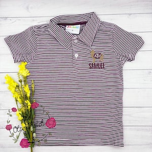 May include: A maroon and white striped polo shirt with a small embroidered llama wearing a maroon and white hat with the letters "ATM" on it. The llama is holding a maroon and white banner with the word "SAMMUELL" on it.