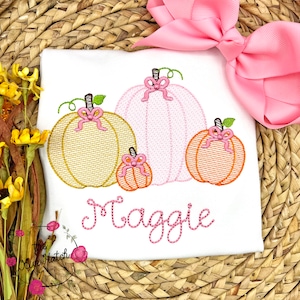 Op de afbeelding: Wit T-shirt met geborduurde pompoenen in goud, roze en oranje, elk versierd met een roze strik. De naam "Maggie" is eronder in roze script geborduurd. Een grote roze strik op de achtergrond.