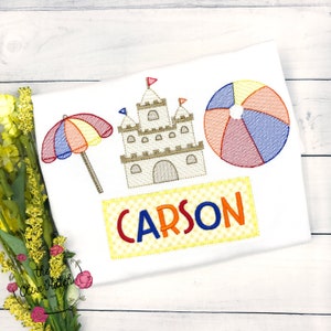 May include: Embroidered design featuring a sandcastle, beach umbrella, beach ball, and the name "CARSON" on a white t-shirt.