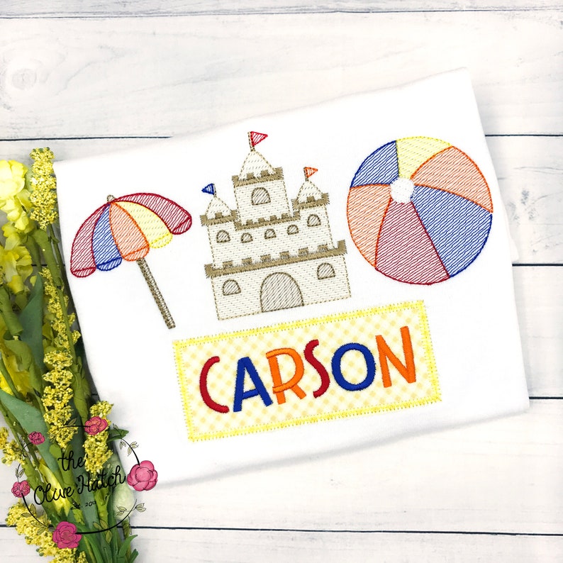 May include: Embroidered design featuring a sandcastle, beach umbrella, beach ball, and the name "CARSON" on a white t-shirt.