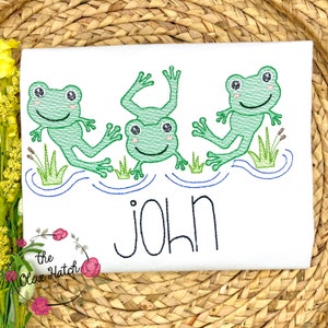 May include: White embroidered t-shirt with three green frogs jumping in a blue pond. The shirt has the name "JOHN" embroidered below the frogs.