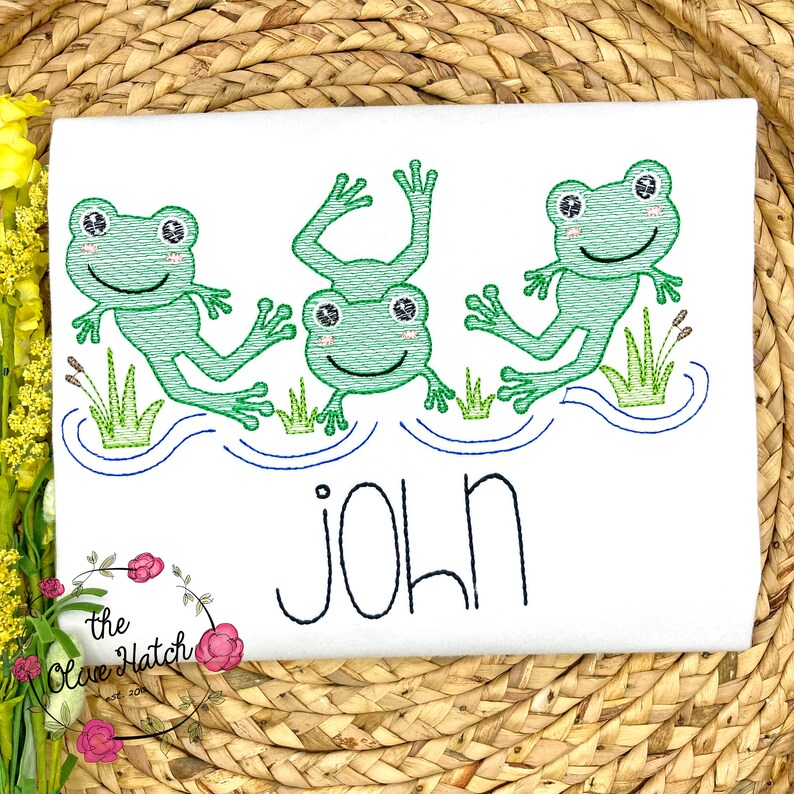 May include: White embroidered t-shirt with three green frogs jumping in a blue pond. The shirt has the name "JOHN" embroidered below the frogs.