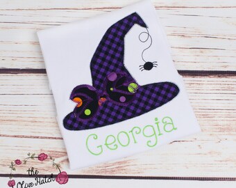 May include: A white t-shirt featuring an embroidered purple and black plaid witch hat applique. The hat is decorated with small orange and lime green circular accents and a dangling black spider. The name Georgia is embroidered in lime green.