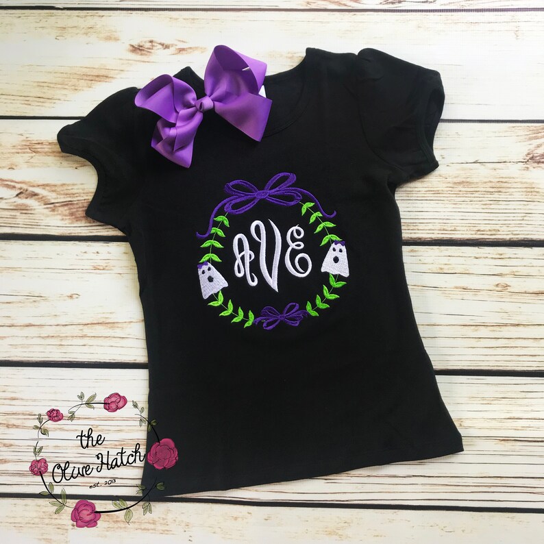 Peut inclure: T-shirt noir &agrave; manches courtes avec un n&oelig;ud violet et une broderie sur le th&egrave;me d'Halloween. Le motif comprend une couronne avec les lettres "AVE", deux fant&ocirc;mes et des n&oelig;uds d&eacute;coratifs. Le logo Olive Hatch est visible.