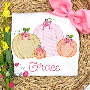 Op de afbeelding: Wit T-shirt met een geborduurd ontwerp van vier pompoenen in goud, roze en oranje, elk versierd met een roze strik. De naam "Grace" is in roze script onder de pompoenen geborduurd. Een grote roze strik staat op de achtergrond.
