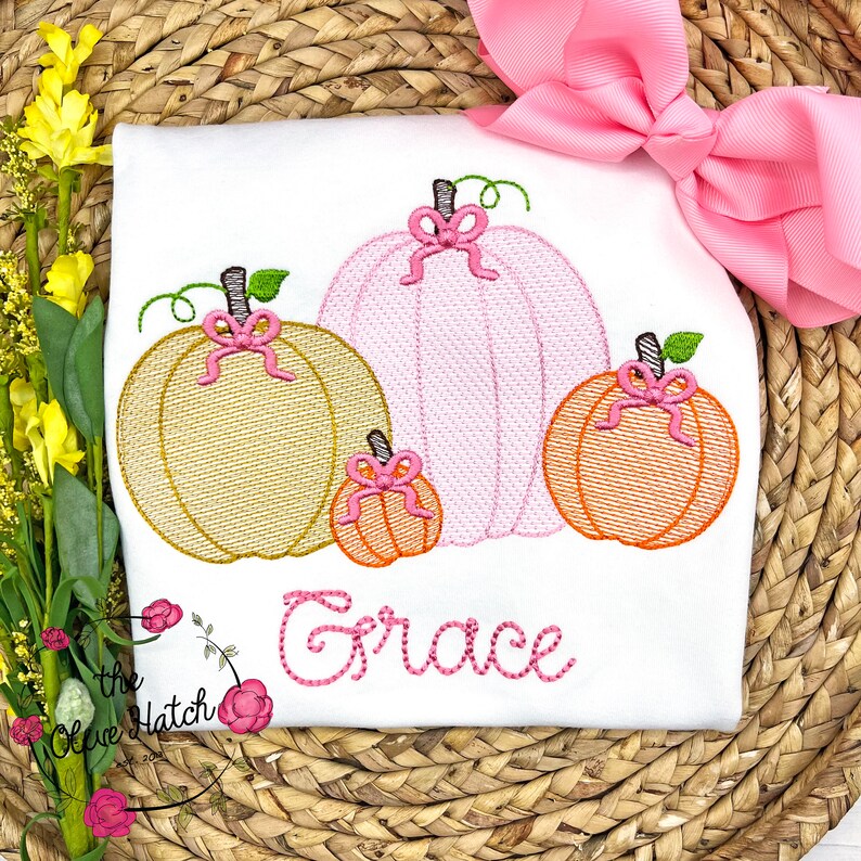 Op de afbeelding: Wit T-shirt met een geborduurd ontwerp van vier pompoenen in goud, roze en oranje, elk versierd met een roze strik. De naam "Grace" is in roze script onder de pompoenen geborduurd. Een grote roze strik staat op de achtergrond.