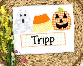 May include: White t-shirt with Halloween-themed appliqués: a ghost, candy corn, and a jack-o'-lantern. The name "Tripp" is embroidered on a green polka-dot rectangle. The shirt is likely for children.