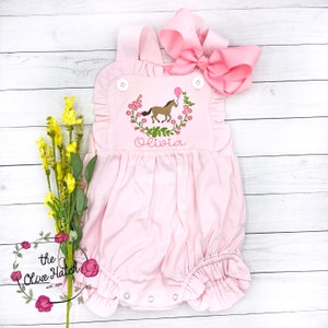 May include: Pink baby onesie with a floral wreath embroidered with a brown horse and a pink balloon. The onesie has a ruffle trim and the name "Olivia" is embroidered below the horse.