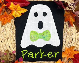 May include: Black t-shirt featuring a white ghost applique with a green bow tie. The ghost has black eyes and an open mouth. The name "Parker" is embroidered in green below the ghost. Autumn leaves surround the shirt.