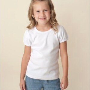 Peut inclure: Une jeune personne porte un t-shirt blanc &agrave; manches courtes et un short en jean. La personne sourit et a les cheveux blonds. Le fond est de couleur beige clair.