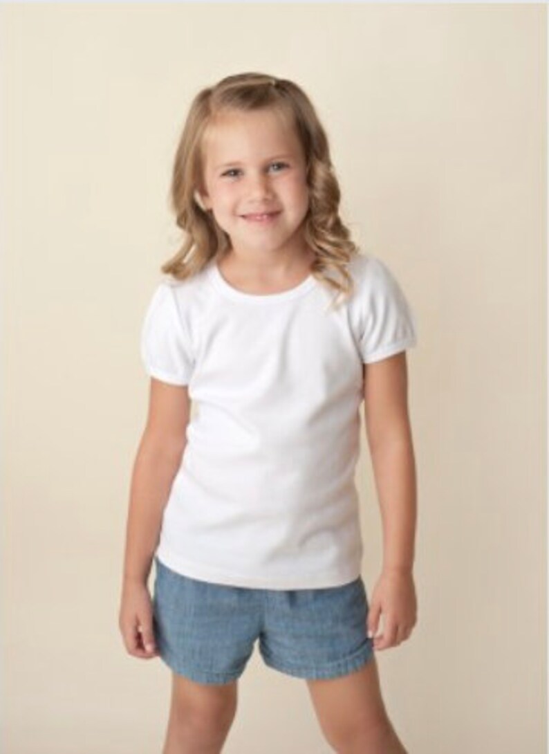 Peut inclure: Une jeune personne porte un t-shirt blanc &agrave; manches courtes et un short en jean. La personne sourit et a les cheveux blonds. Le fond est de couleur beige clair.