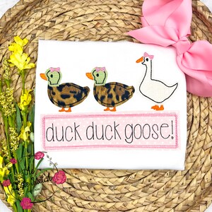 Op de afbeelding: Wit T-shirt met een geborduurd ontwerp van twee eenden en een gans met de tekst "duck duck goose!"