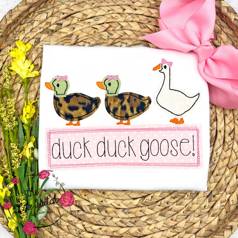 Op de afbeelding: Wit T-shirt met een geborduurd ontwerp van twee eenden en een gans met de tekst "duck duck goose!"