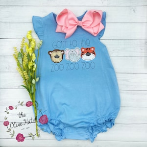 May include: A light blue baby onesie with a ruffle trim. The onesie has a graphic of three animal faces with the text "goin' to the zoo zoo zoo". A pink bow is on top of the onesie.