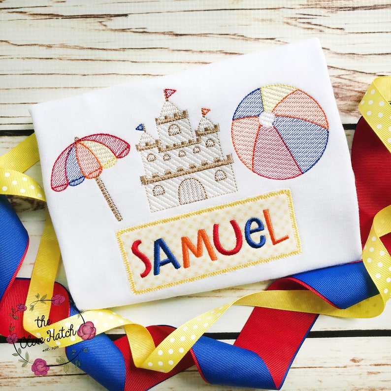 May include: A white embroidered t-shirt with a beach theme. The design includes a sandcastle, beach umbrella, beach ball, and the name "SAMUEL".