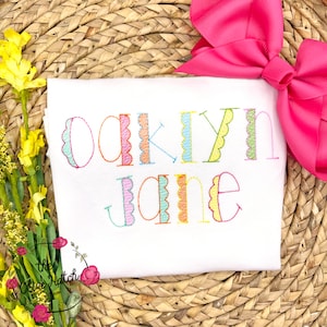 May include: White t-shirt with the name "Oaklyn Jane" embroidered in colorful thread. The letters have a scalloped edge design. A large pink bow is in the upper right corner, and yellow flowers are on the left side.