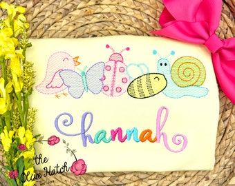 May include: A light yellow embroidered shirt with a bird, butterfly, ladybug, bee, and snail design. The shirt has the name "Hannah" embroidered in colorful letters.