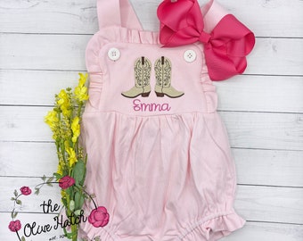 Könnte beinhalten: Rosaer Baby-Strampler mit Rüschen am Ausschnitt und Trägern. Der Strampler hat ein gesticktes Design mit zwei braunen Cowboystiefeln und dem Namen "Emma". Eine rosa Schleife ist im Lieferumfang enthalten.