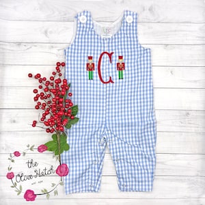 May include: Blue and white gingham baby romper with a red embroidered letter "C" and two toy soldier designs. The romper has white buttons and snaps. The Olive Hatch logo is in the bottom left corner.