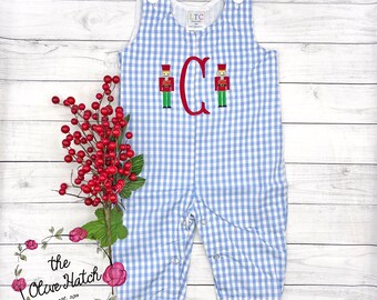 May include: Baby romper in blue and white gingham, featuring a red embroidered letter "C" and two toy soldier designs. It has white buttons and poppers. The Olive Hatch logo is in the bottom left corner.