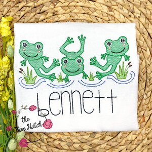 May include: White t-shirt with an embroidered design of three green frogs jumping in a blue pond. The name "Bennett" is embroidered below the frogs.