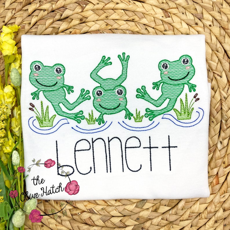 May include: White t-shirt with an embroidered design of three green frogs jumping in a blue pond. The name "Bennett" is embroidered below the frogs.