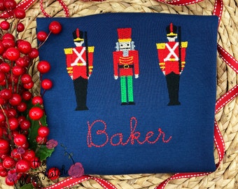 May include: Navy blue shirt with embroidered toy soldier and nutcracker designs in red, black, and gold. The name "Baker" is embroidered in red script. The shirt is surrounded by red berries and a red and white ribbon.