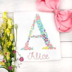 May include: A white onesie with a floral embroidered letter A and the name "Alice" embroidered below it. The onesie is on a white wooden surface with a pink bow and a bouquet of yellow flowers.