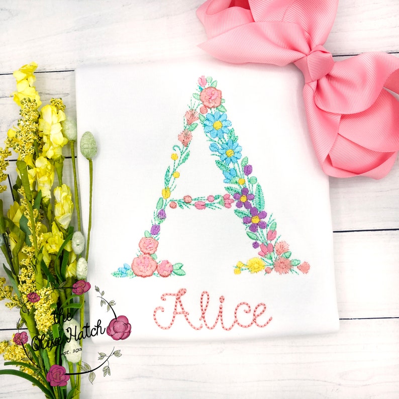 May include: A white onesie with a floral embroidered letter A and the name "Alice" embroidered below it. The onesie is on a white wooden surface with a pink bow and a bouquet of yellow flowers.