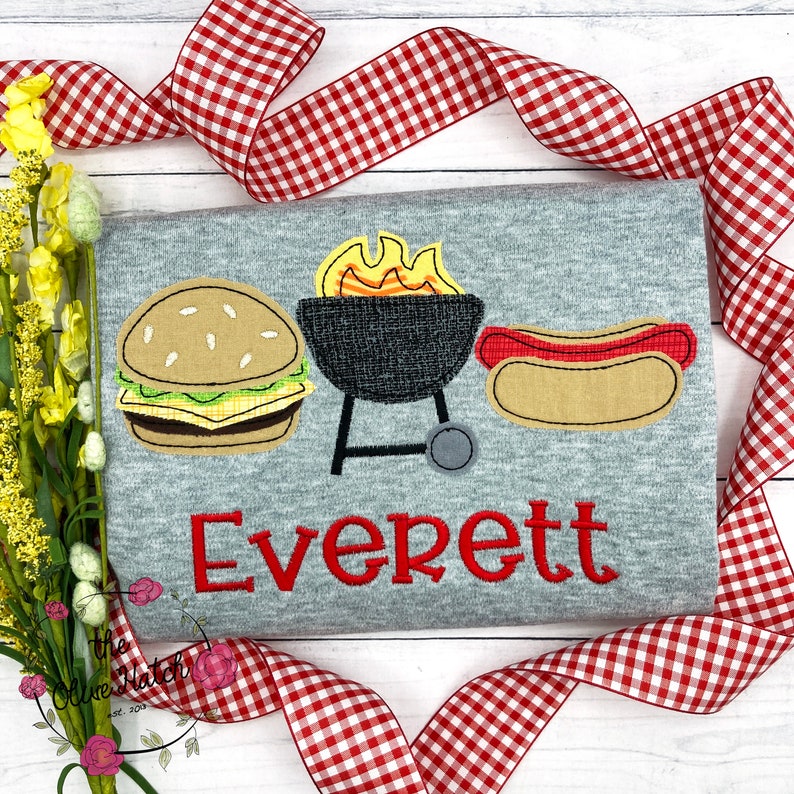 May include: A gray t-shirt with a red embroidered name "Everett" and a design of a hamburger, hot dog, and grill with flames.