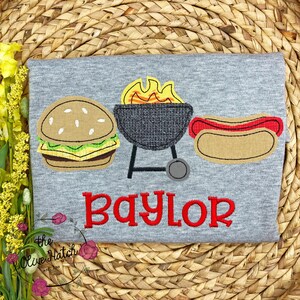 May include: Gray t-shirt with an embroidered design of a hamburger, a grill with flames, and a hot dog. The text "Baylor" is embroidered in red below the design.