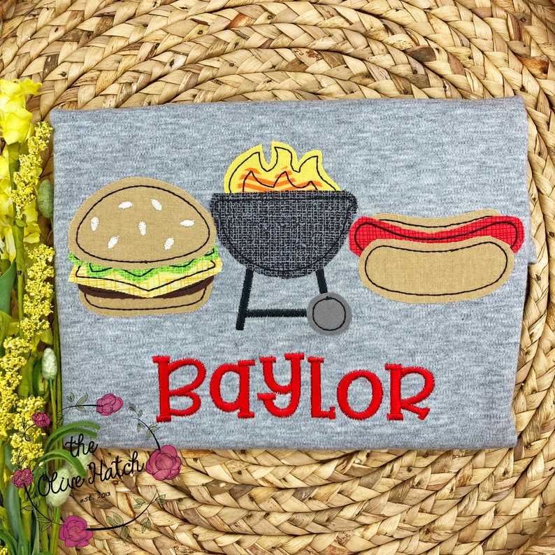 May include: Gray t-shirt with an embroidered design of a hamburger, a grill with flames, and a hot dog. The text "Baylor" is embroidered in red below the design.