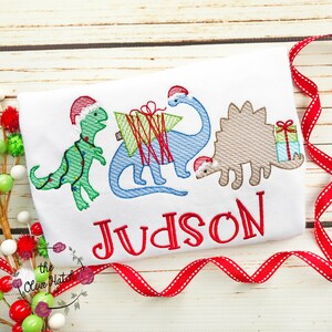 May include: White t-shirt featuring embroidered Christmas dinosaurs wearing Santa hats. The dinosaurs are green, blue, and tan, with a red embroidered name "Judson" below. A red ribbon and decorative ornaments are in the background.