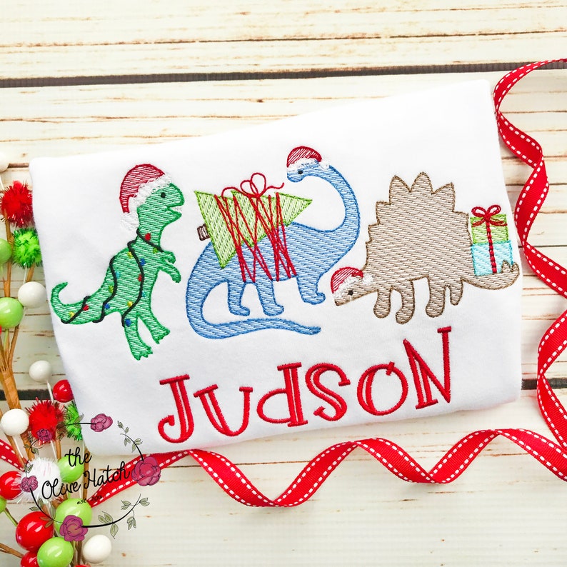 May include: White t-shirt featuring embroidered Christmas dinosaurs wearing Santa hats. The dinosaurs are green, blue, and tan, with a red embroidered name "Judson" below. A red ribbon and decorative ornaments are in the background.
