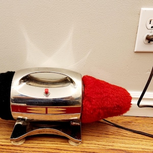 Op de afbeelding: Een vintage elektrische schoenpoetsmachine met een zwarte en rode borstel. De poetsmachine is zilverkleurig met een rode knop en een gouden streep. Hij is aangesloten op een stopcontact.