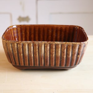 May include: A brown and gold ceramic planter with a ribbed design. The planter is square shaped and has a wide opening.