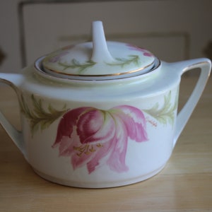 May include: White porcelain sugar bowl with a lid, featuring a floral design of pink and green tulips. The bowl has a gold trim around the lid and base.