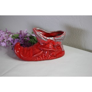May include: Red ceramic shoe planter with white glaze. The shoe has a pointed toe and a raised heel. The planter is decorated with a mushroom design.