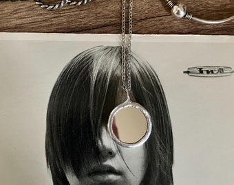 May include: A silver necklace with a round pendant, a silver bracelet, and twisted silver hoop earrings are displayed on a wooden surface. The pendant has a reflective surface. A black and white photo of a person is in the background.