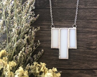 May include: A silver chain necklace with a pendant of three white rectangular glass panels. The pendant is set in a silver metal frame. The background is a dark wood surface, with dried flowers on the left.