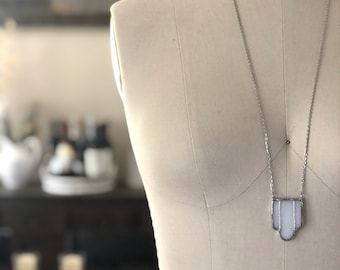 May include: A silver necklace with a white pendant is displayed on a mannequin. The pendant has a geometric design with a white centre and a silver border. The background is blurred, but includes the word "Leatherlily".