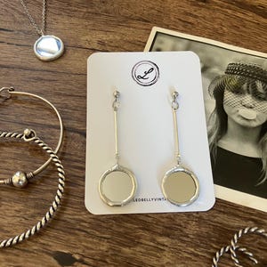 Puede incluir: Juego de joyas plateadas sobre una superficie de madera. El conjunto incluye un collar con colgante redondo, pendientes largos con detalles circulares espejados y varias pulseras. Una fotografía en blanco y negro se encuentra en el fondo.